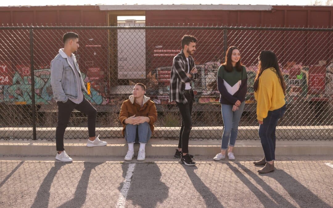 Five young people hang out in a sunlit car park beside a chain-link fence, with a graffiti-covered freight train behind them; one sits on the kerb while the others stand chatting, casting long shadows.